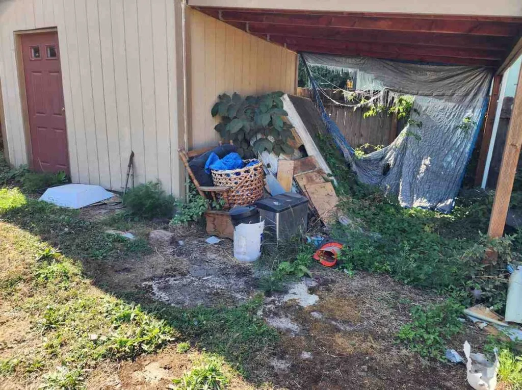 Cluttered side yard under a carport in Gresham with abandoned furniture, old tarps, and overgrown vegetation requiring professional hauling.