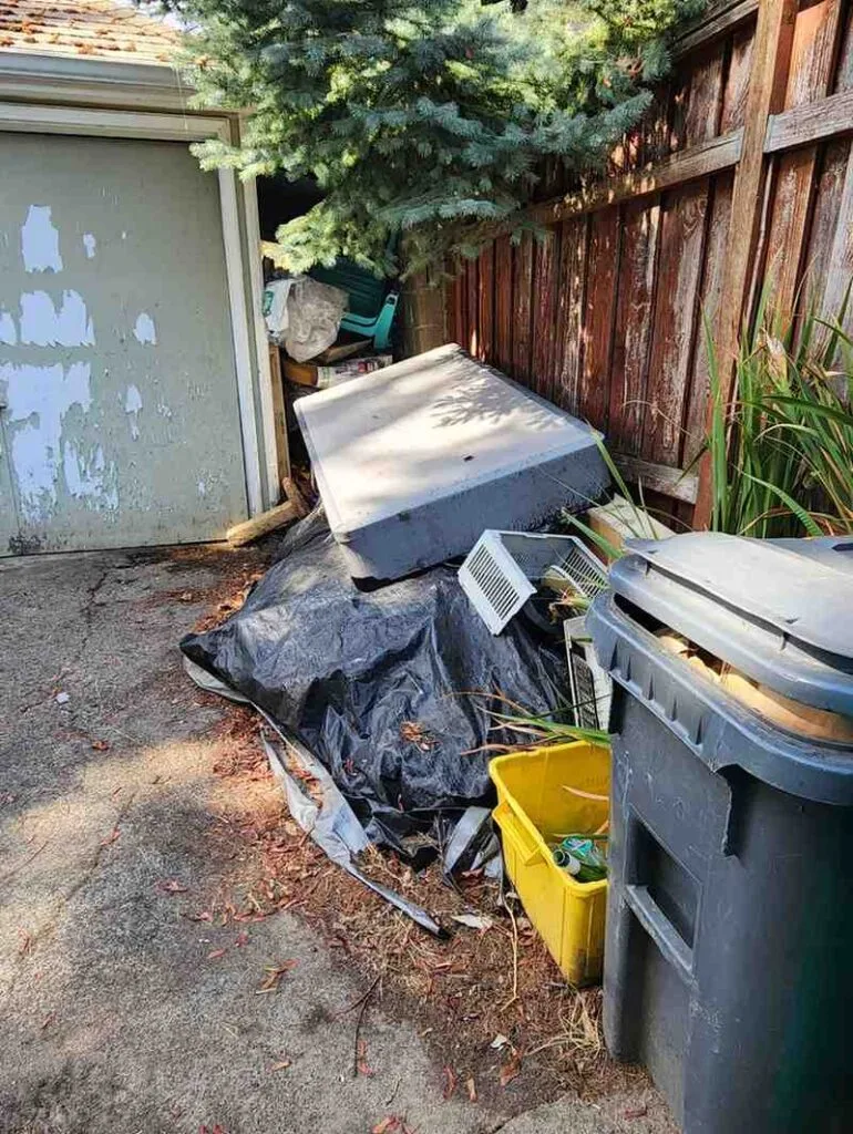 Outdoor utility area near an HVAC unit cluttered with old wooden furniture pieces, metal frames, and scattered debris.