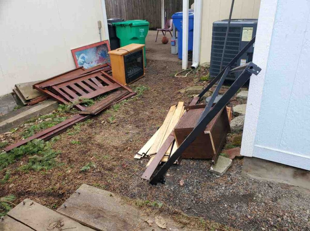 Narrow walkway between a garage and fence piled with a mattress, black plastic bags, and miscellaneous household waste.