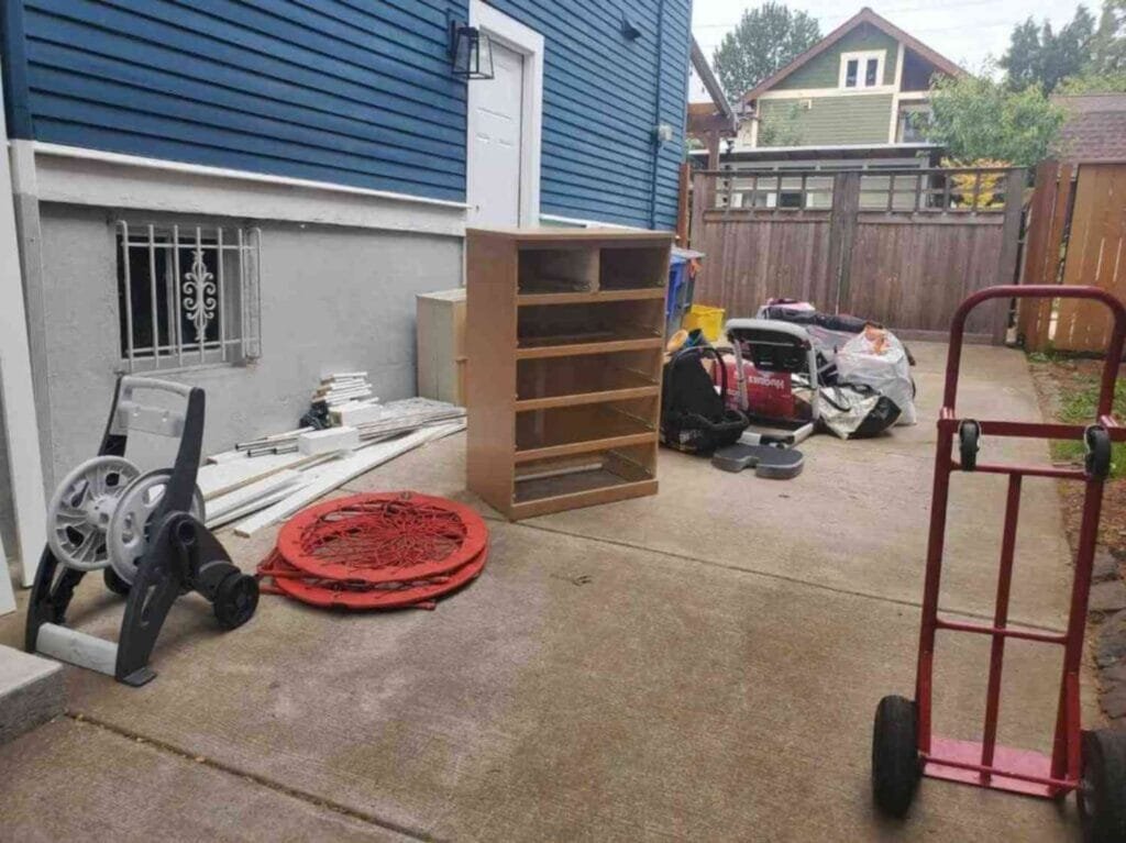 Cluttered concrete driveway in Portland filled with old furniture, a dresser, baby gear, and loose debris before junk removal.