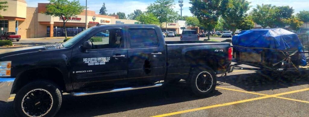 503 Clutter Busters LLC black Chevy 2500 HD 4x4 truck with branded door lettering and trailer loaded with junk covered by blue tarp - professional junk removal service Portland Oregon